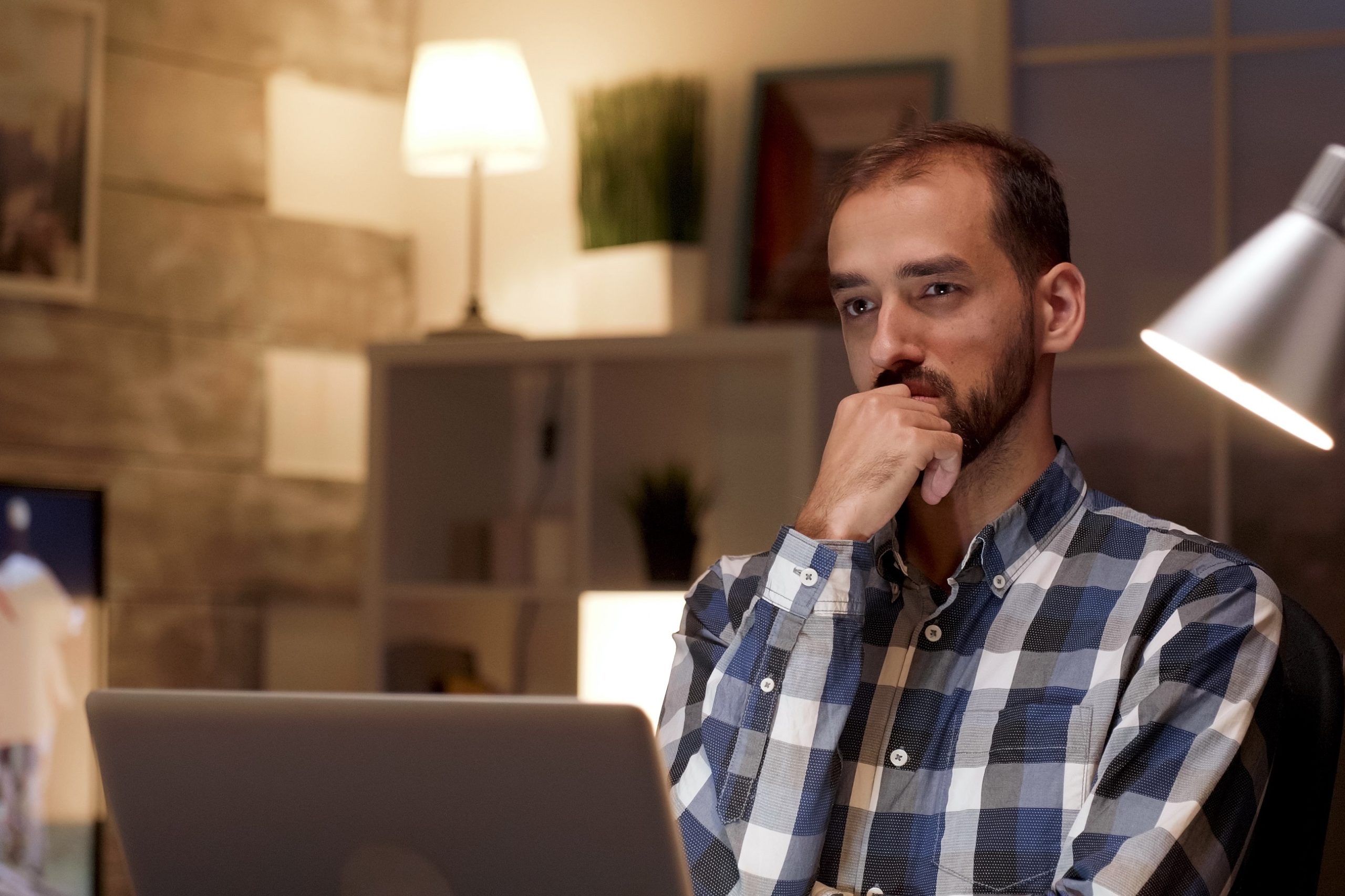 Businessman thinking and holding arms crossed while working on laptop in home office during night hours.